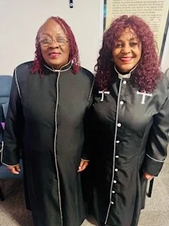 Two women standing side by side, smiling, wearing black and gray religious robes with white crosses on the chest, in an indoor setting.