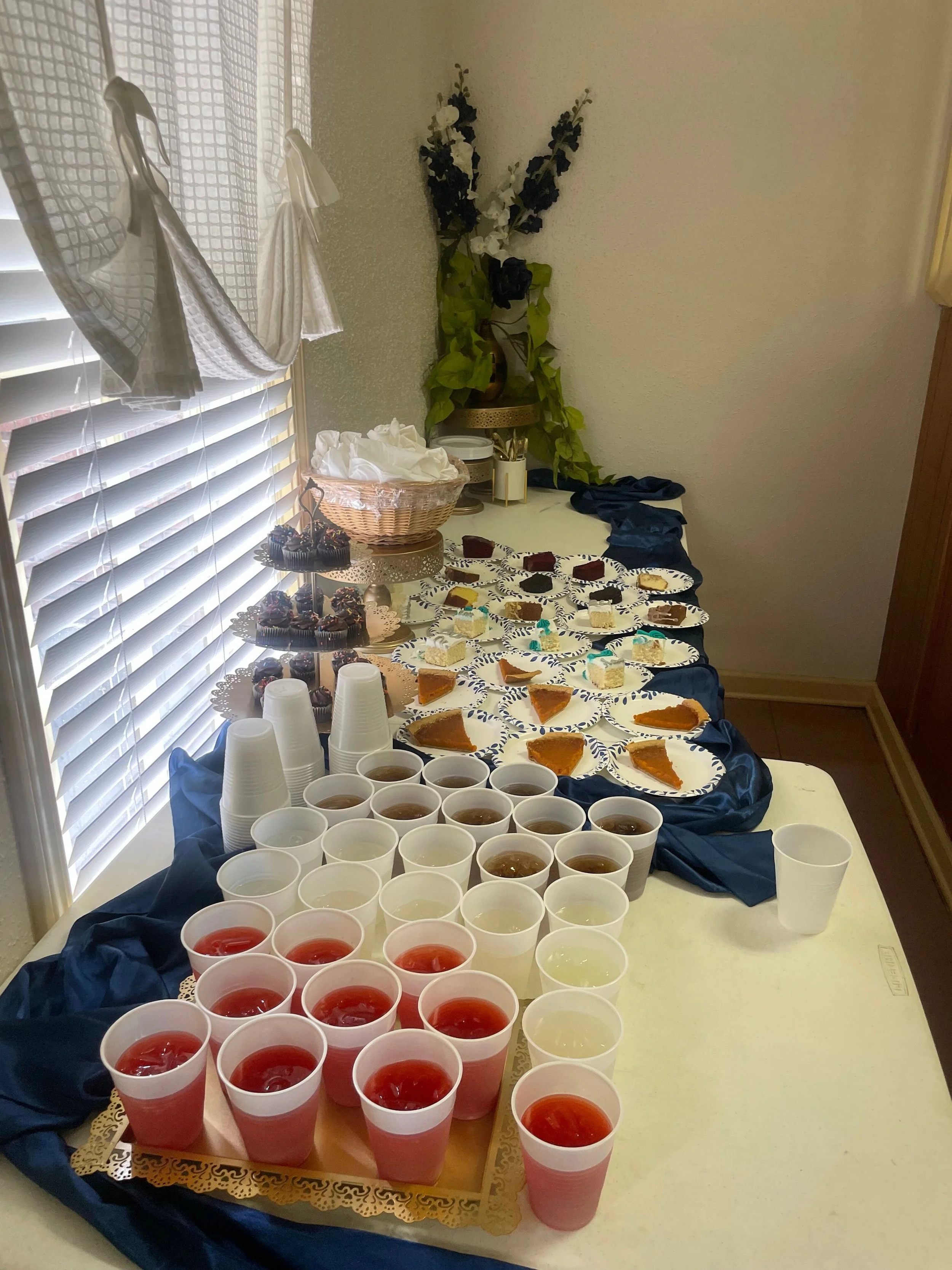Table set with drinks, desserts, and snacks for a party or gathering, including red and white punch, various cakes, and cupcakes, with a decorative arrangement in the background.