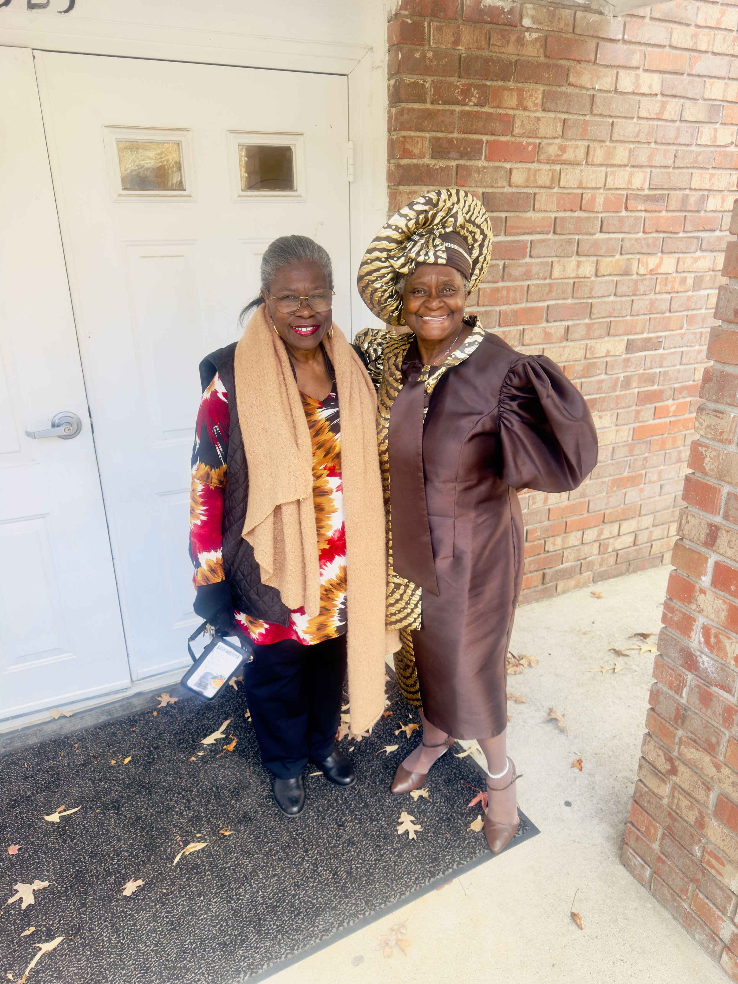 Two women standing outdoors in front of a brick wall and a white door, smiling at the camera. One woman is dressed in a colorful patterned top, black pants, and a tan scarf. The other woman is wearing traditional African attire with a headwrap.