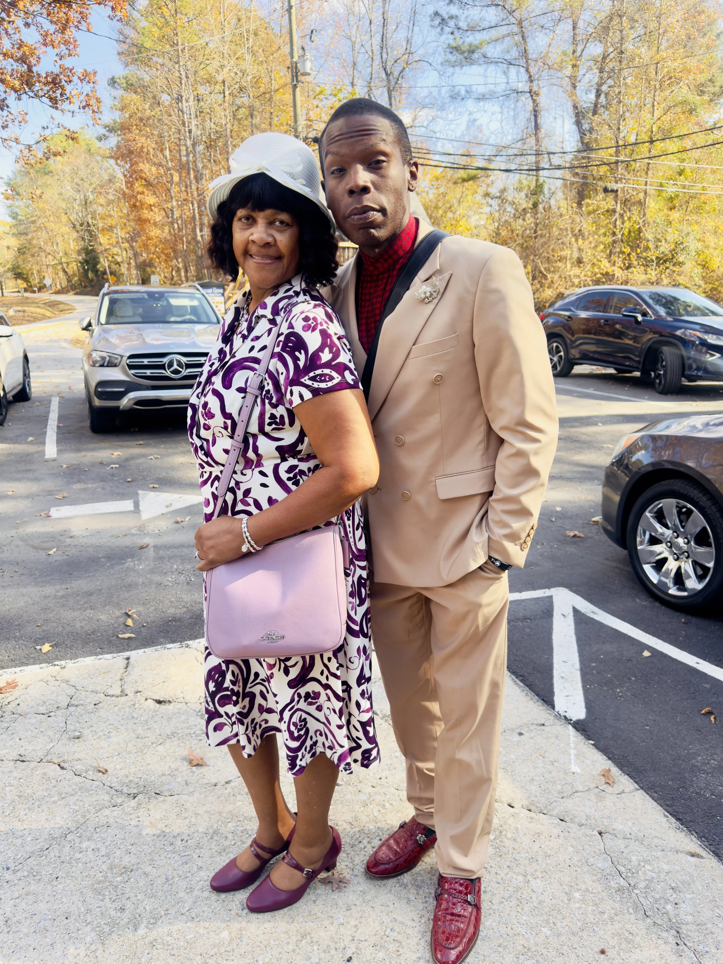 A woman and a man standing on a parking lot, posing for a photo. The woman is wearing a purple and white patterned dress, a white hat, and purple shoes, and carrying a light purple purse. The man is dressed in a beige suit with a red checkered shirt,