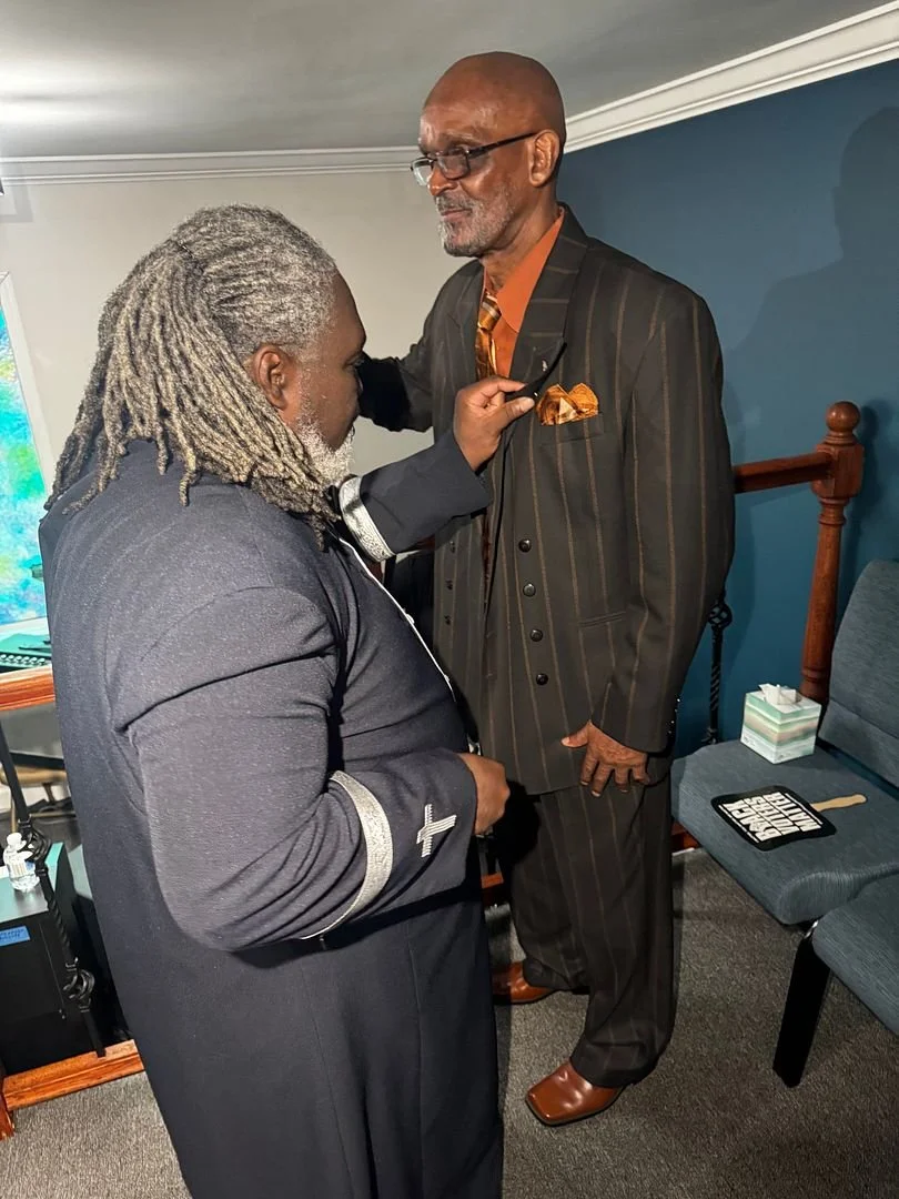 A man in a dark suit and glasses is helping another man in a dark priestly robe adjust his pocket square during a church service or ceremony inside a room with blue walls and chairs.