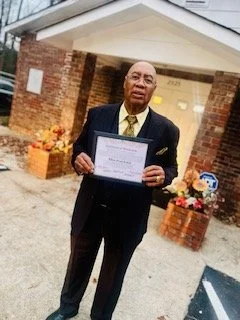 An elderly man in a suit holding a framed certificate outside a house decorated with flowers.