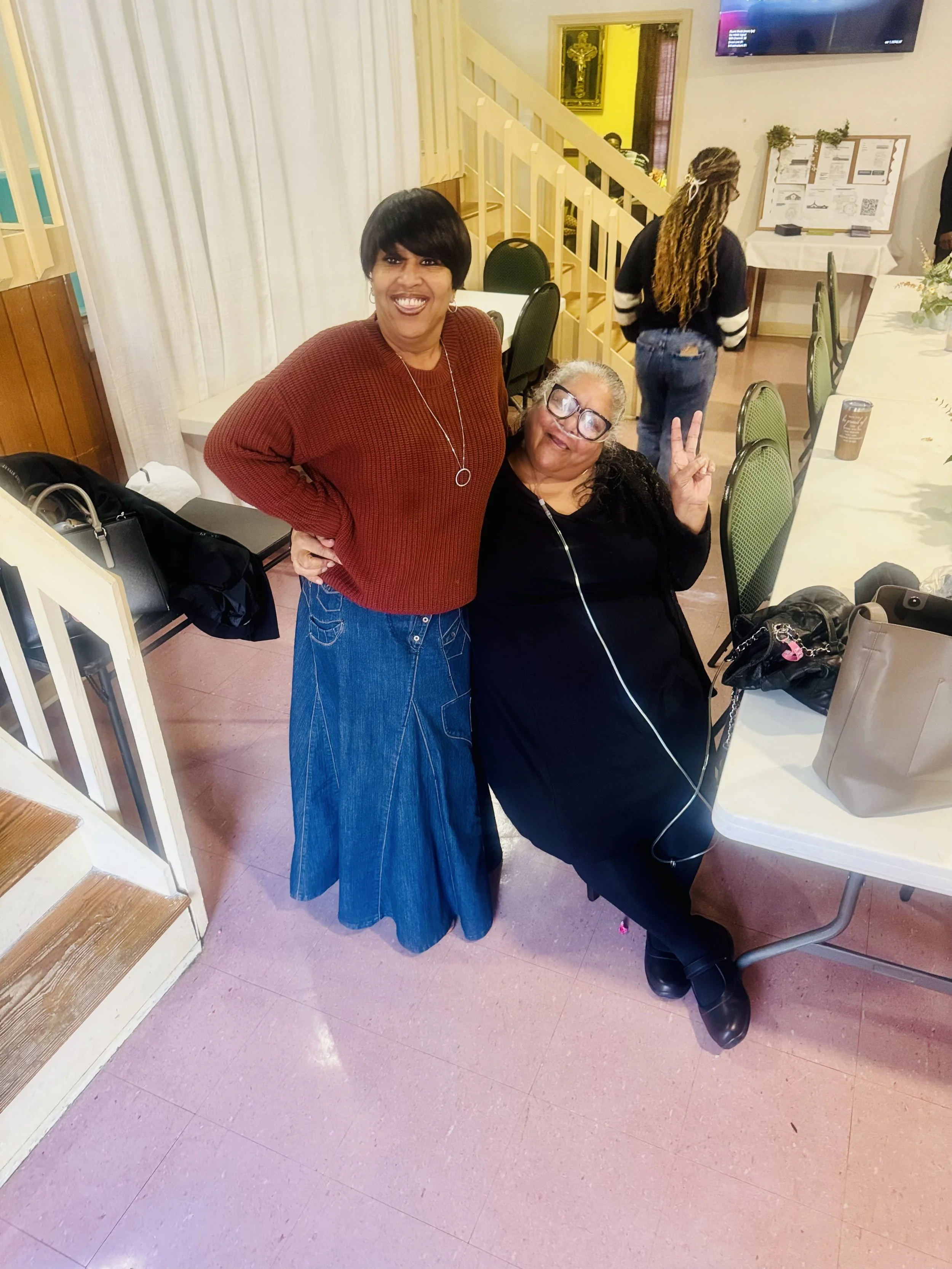 Two women smiling and posing for a photo inside a room. One woman is standing with her arm around the other woman, who is seated and making a peace sign with her fingers. The table nearby has a handbag and some personal items. Background includes a s
