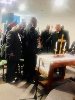 Group of men dressed in suits bowing their heads in prayer around a church altar with a cross and candles.