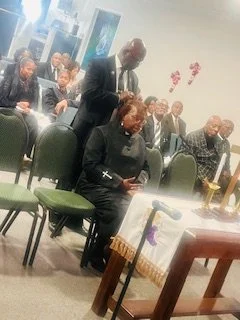 A man in a suit is standing and placing a hand on a woman's shoulder who is seated. The woman is smiling and wearing a dark dress. Several other people are seated in the background, some looking forward and others engaged in conversation, in a room w