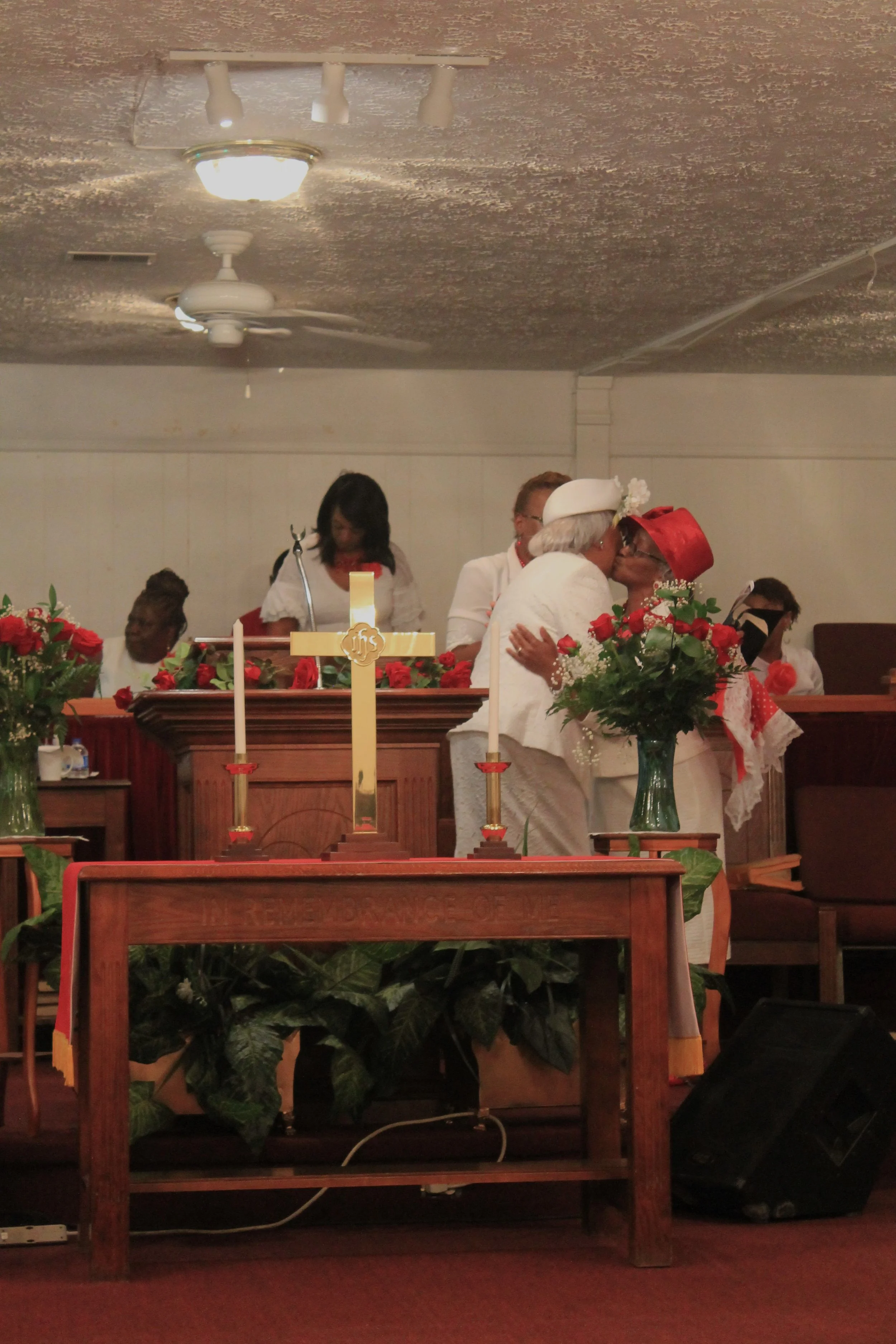 Two women dressed in white and wearing red hats sharing a kiss in front of a church altar decorated with red flowers and candle holders, with other people in white in the background.