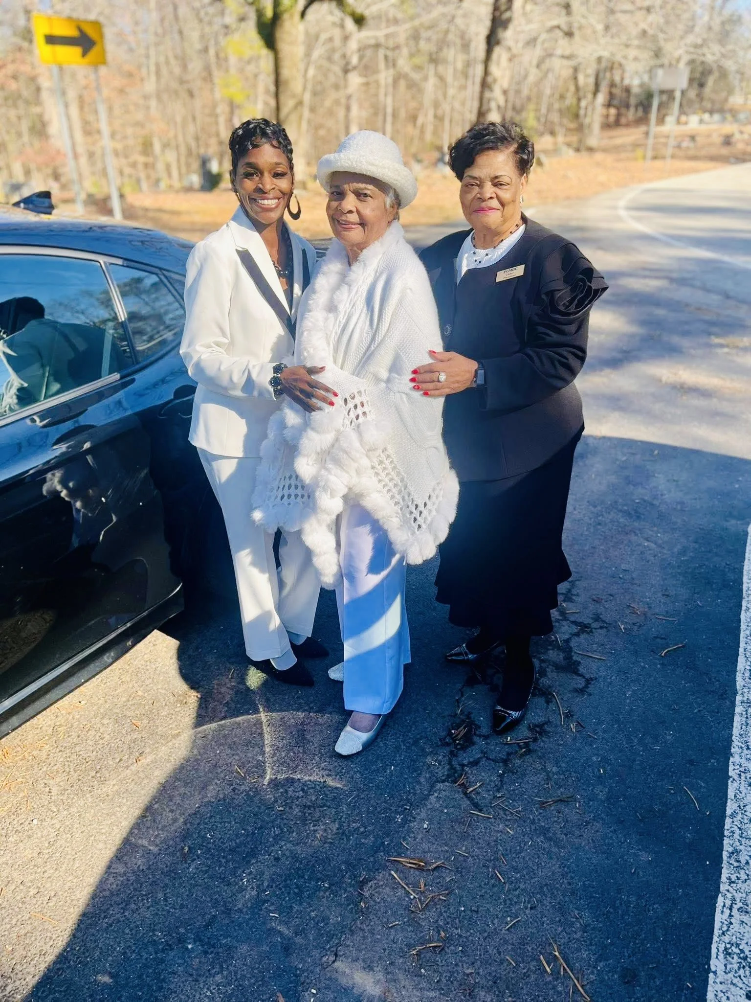 Three women standing outdoors near a black car on a paved surface, surrounded by trees with autumn leaves. They are dressed in elegant clothing, smiling, with one woman in the center wearing white and the other two women on either side wearing black 