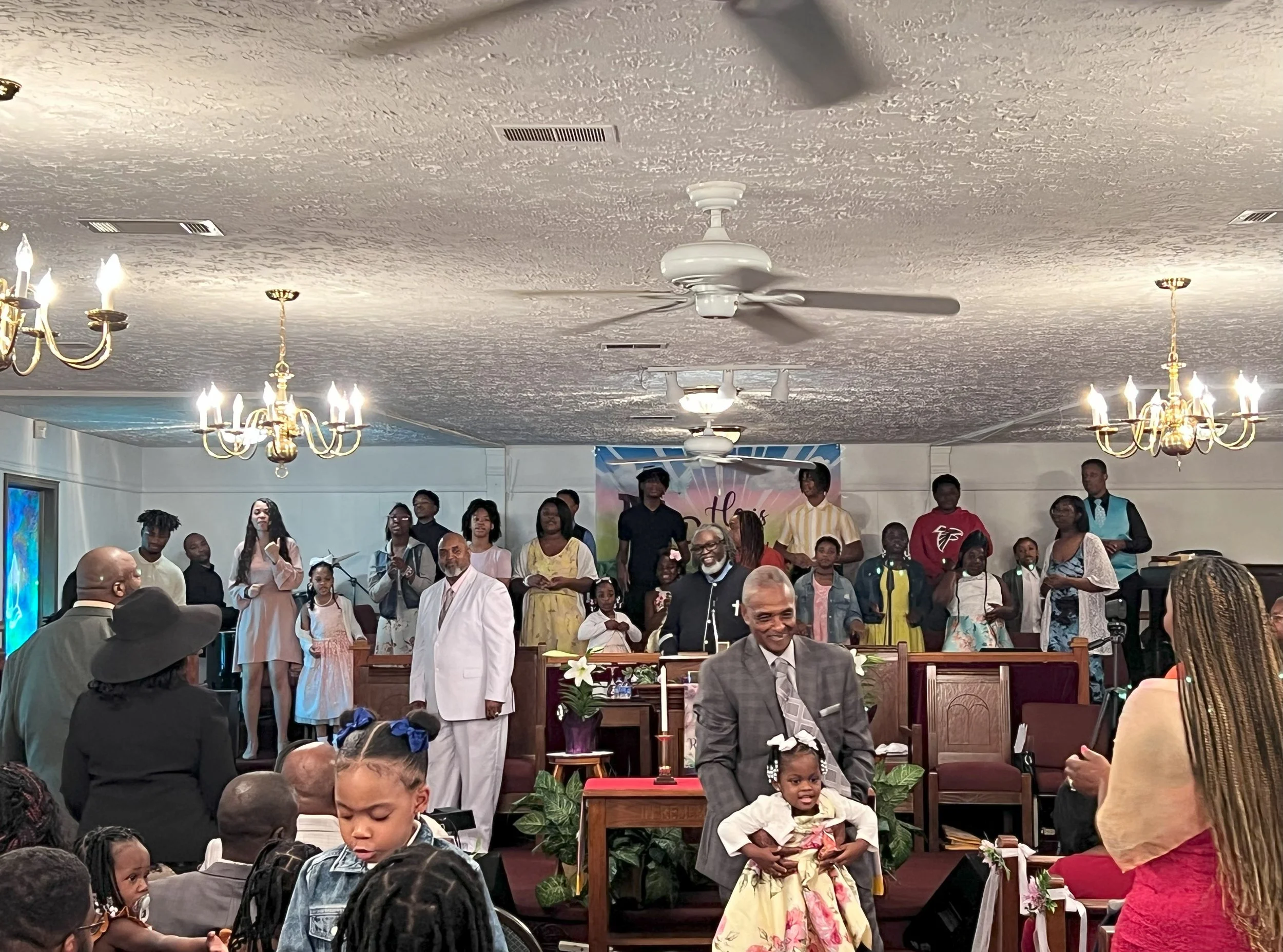 People gathered in a church during a ceremony, with a choir on stage and children in the congregation, some dressed in formal and colorful attire.