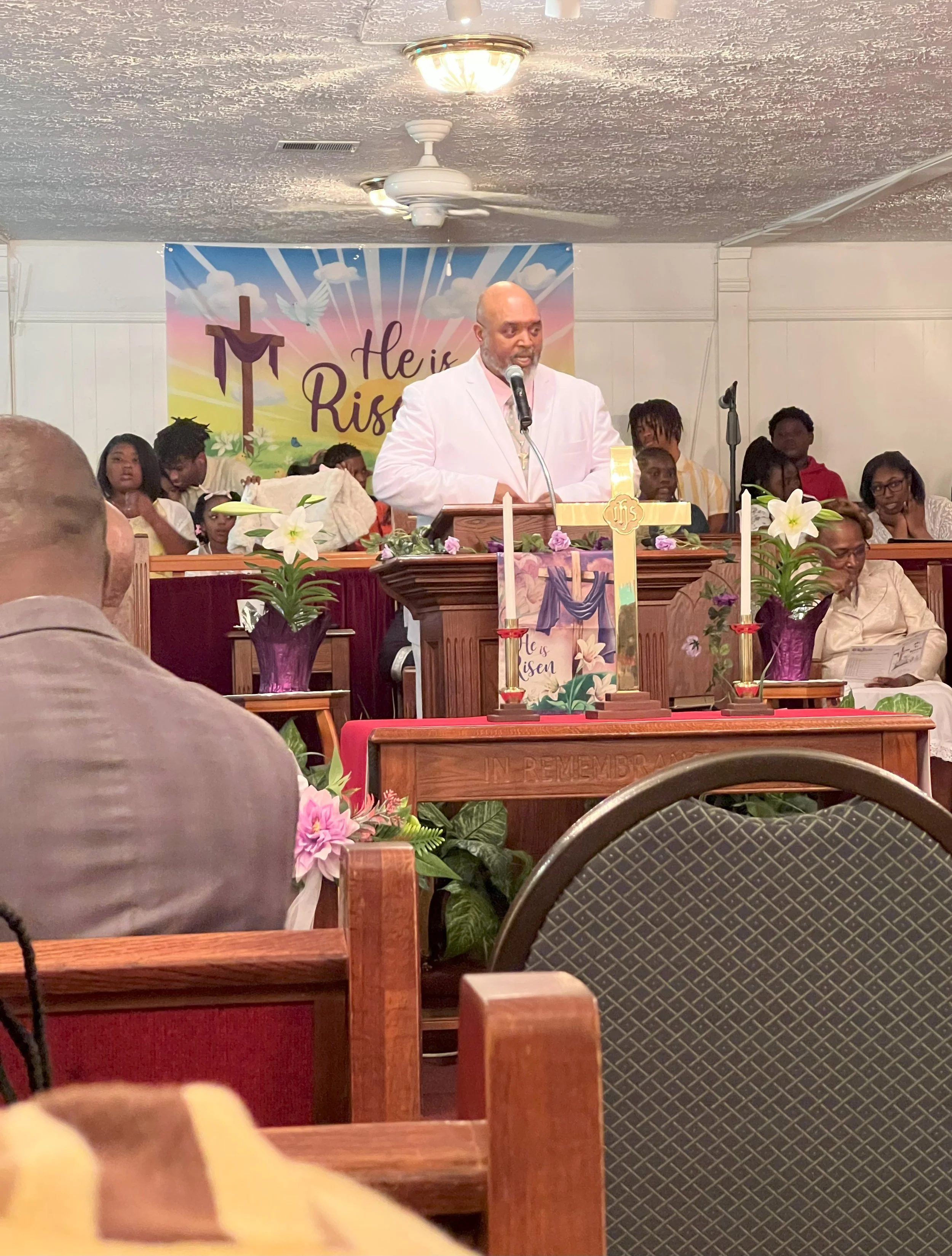 A man in a white suit is speaking at a church service, standing behind a wooden pulpit decorated with flowers and a cross. The background features a large banner with the words 'He is Risen' and religious symbols. Several people are seated behind him