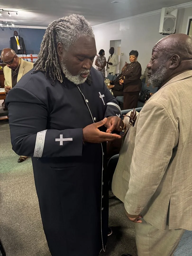 Two elderly men with gray hair and beards are engaged in a conversation in a room with several other people in the background. One man is wearing a black garment with silver cross symbols, while the other is dressed in a light tan suit.