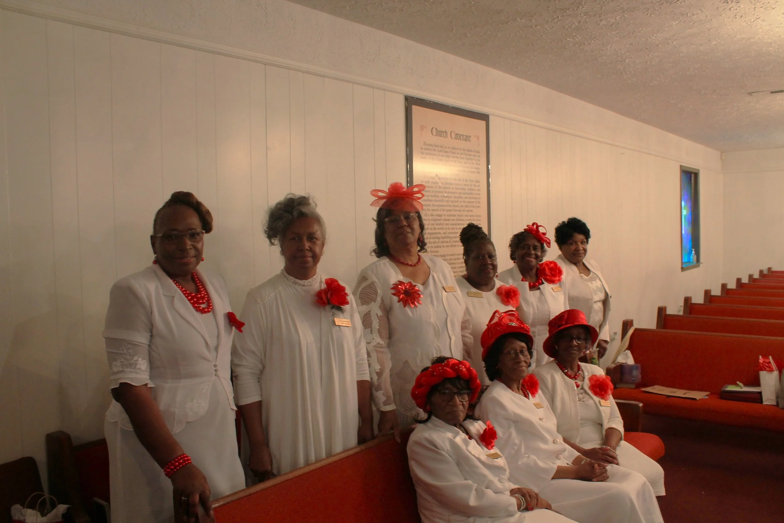 A group of eleven women dressed in white with red accessories, seated and standing in a church pew inside a church, with a framed text on the wall behind them and stained glass windows on the side.