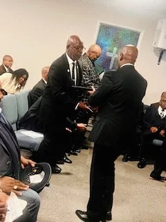 A group of men and women in formal attire gathered in a room for a ceremony or event, with two men in the center exchanging something, possibly a handshake or an award, while others are seated and observing.