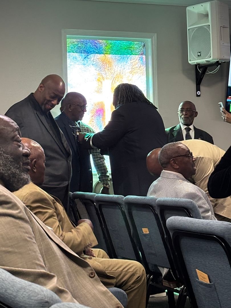 Group of men in formal attire gathered in a room with a stained glass window, some standing and some seated, during a ceremony or event.