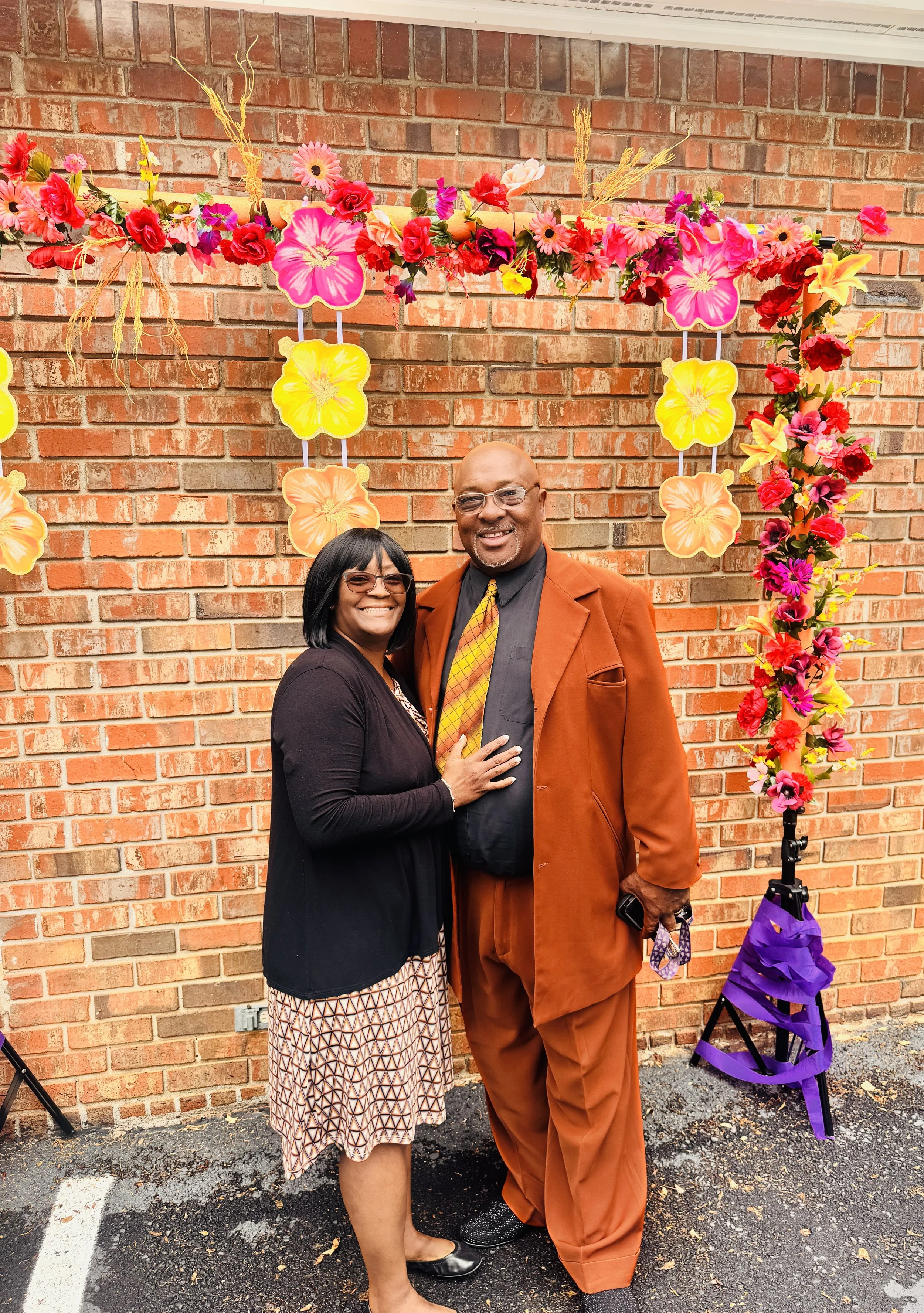 A smiling woman and man stand together in front of a brick wall decorated with a colorful floral arch, wearing semi-formal attire.