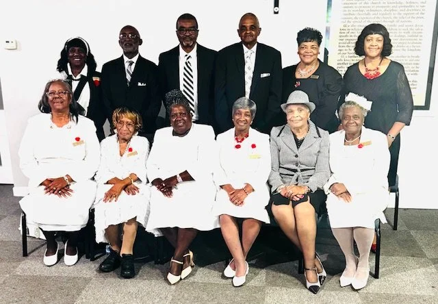 Group of eleven diverse professionals, mostly women, posing for a photo in a formal setting. Some are seated, others are standing, and they are dressed in professional attire, with some women in white coats. The background includes a white wall and a