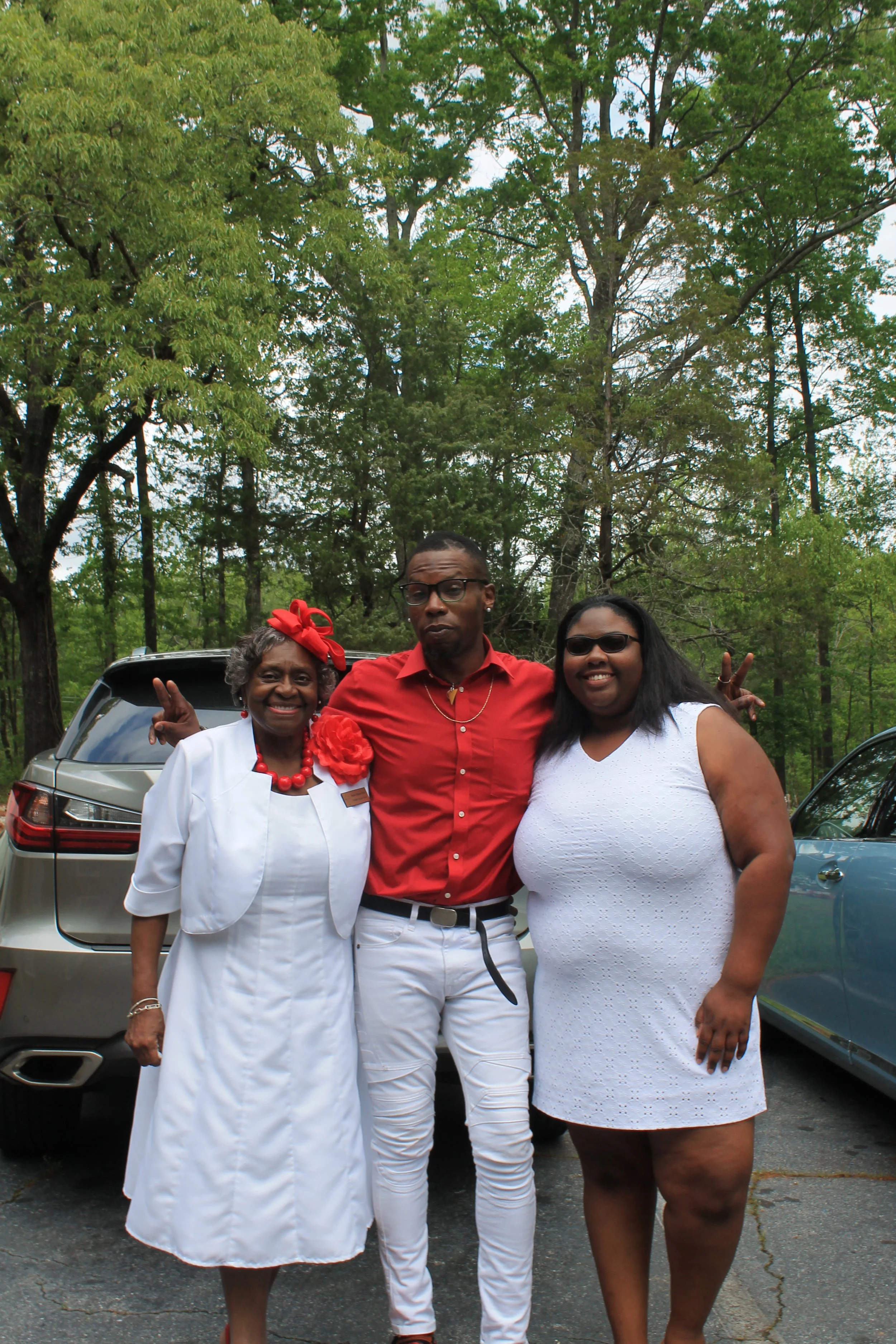 Three people standing outdoors in front of cars and trees, smiling and posing for the photo. The woman on the left is dressed in white with red accessories, the man in the middle is wearing a red shirt and white pants, and the woman on the right is i