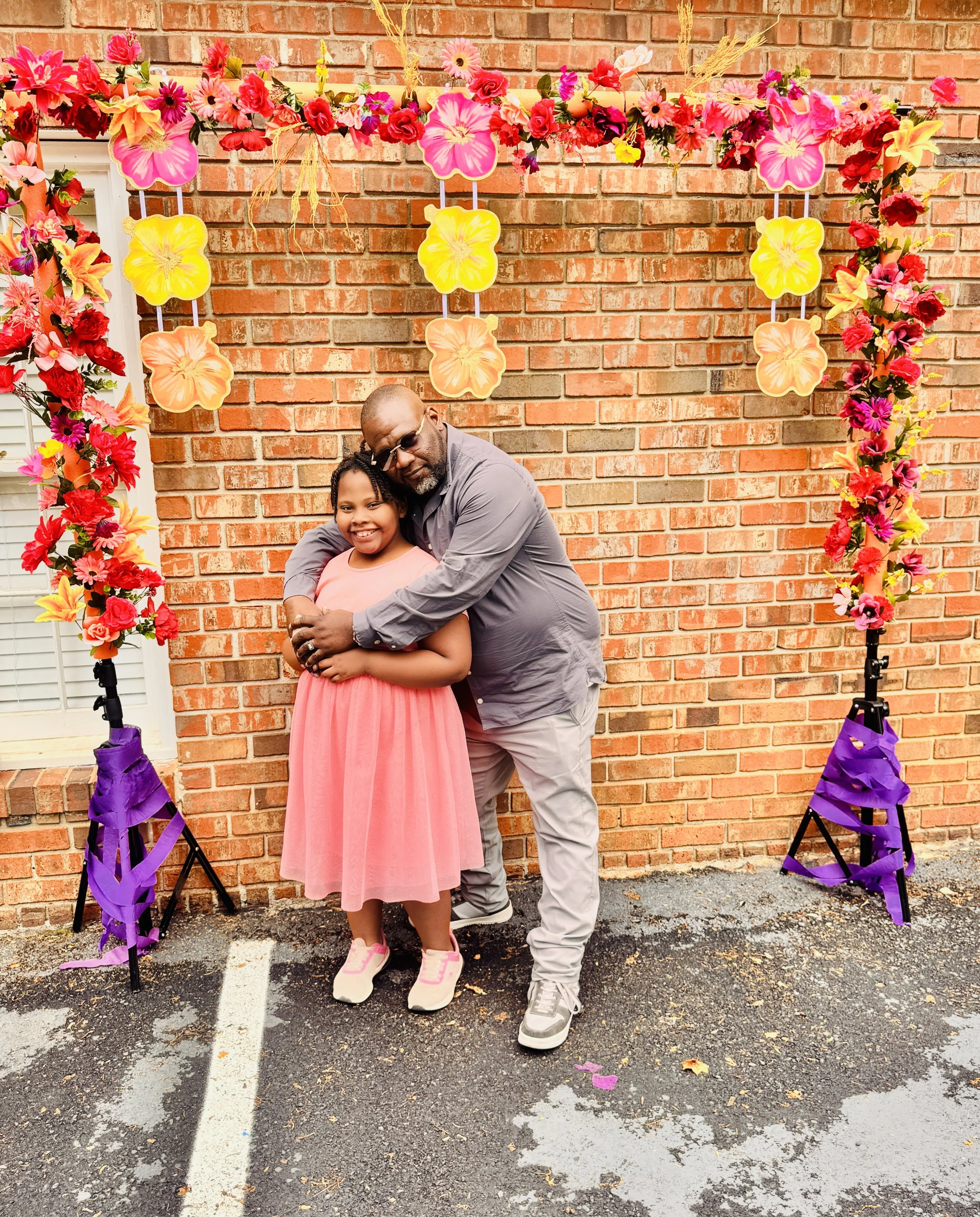 A man and young girl hugging in front of a floral arch with pink, yellow, and orange flowers, set against a brick wall, during a celebration or event.