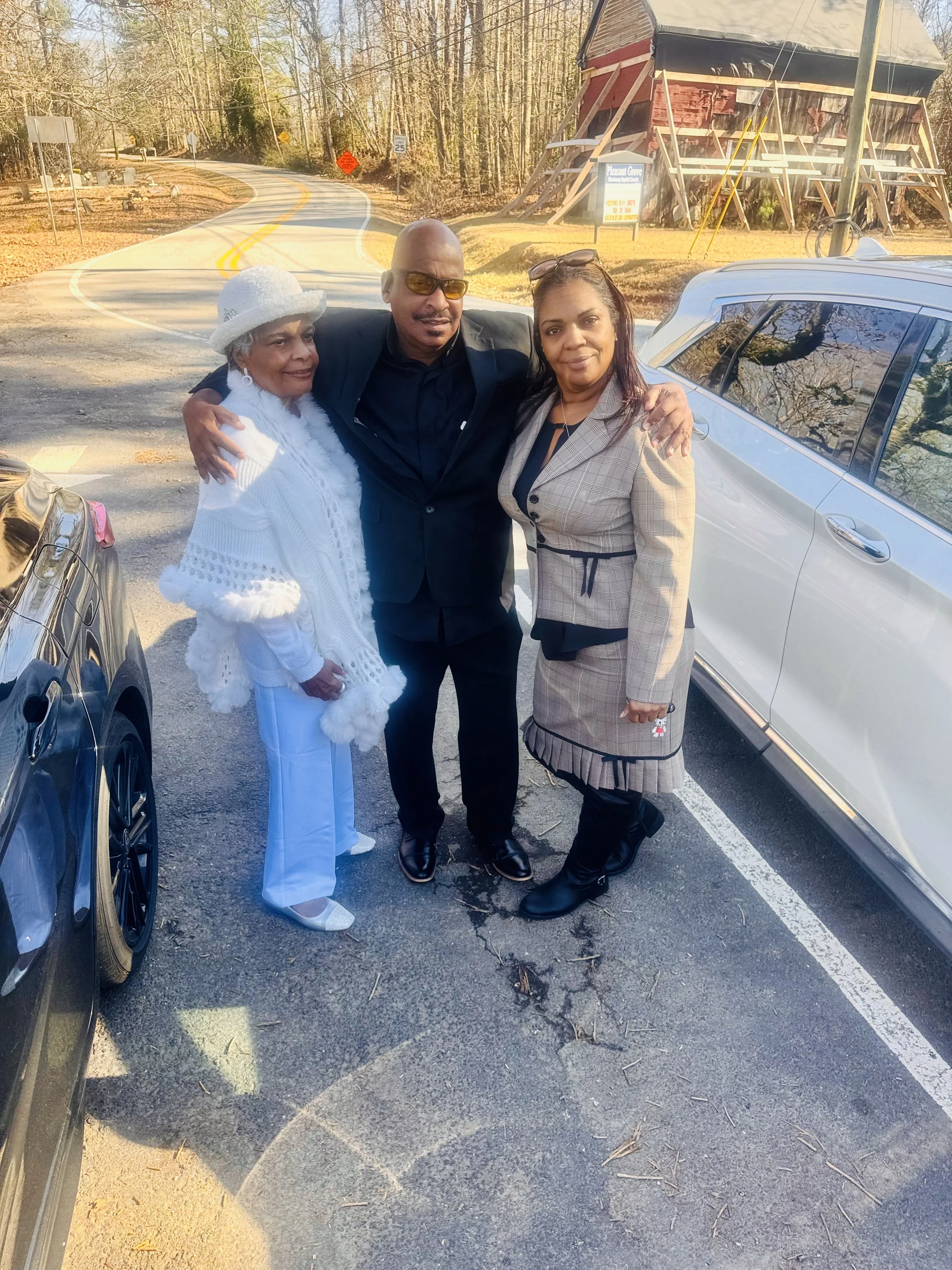 Three people standing together outdoors, smiling, with cars around them and a rustic building in the background.