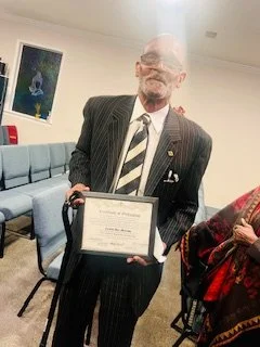 An elderly man in a pinstripe suit holding a framed certificate, standing in a room with blue seating and a framed picture on the wall.