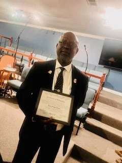 A man in a suit holding a framed certificate in a room with chairs and a large television screen on the wall.