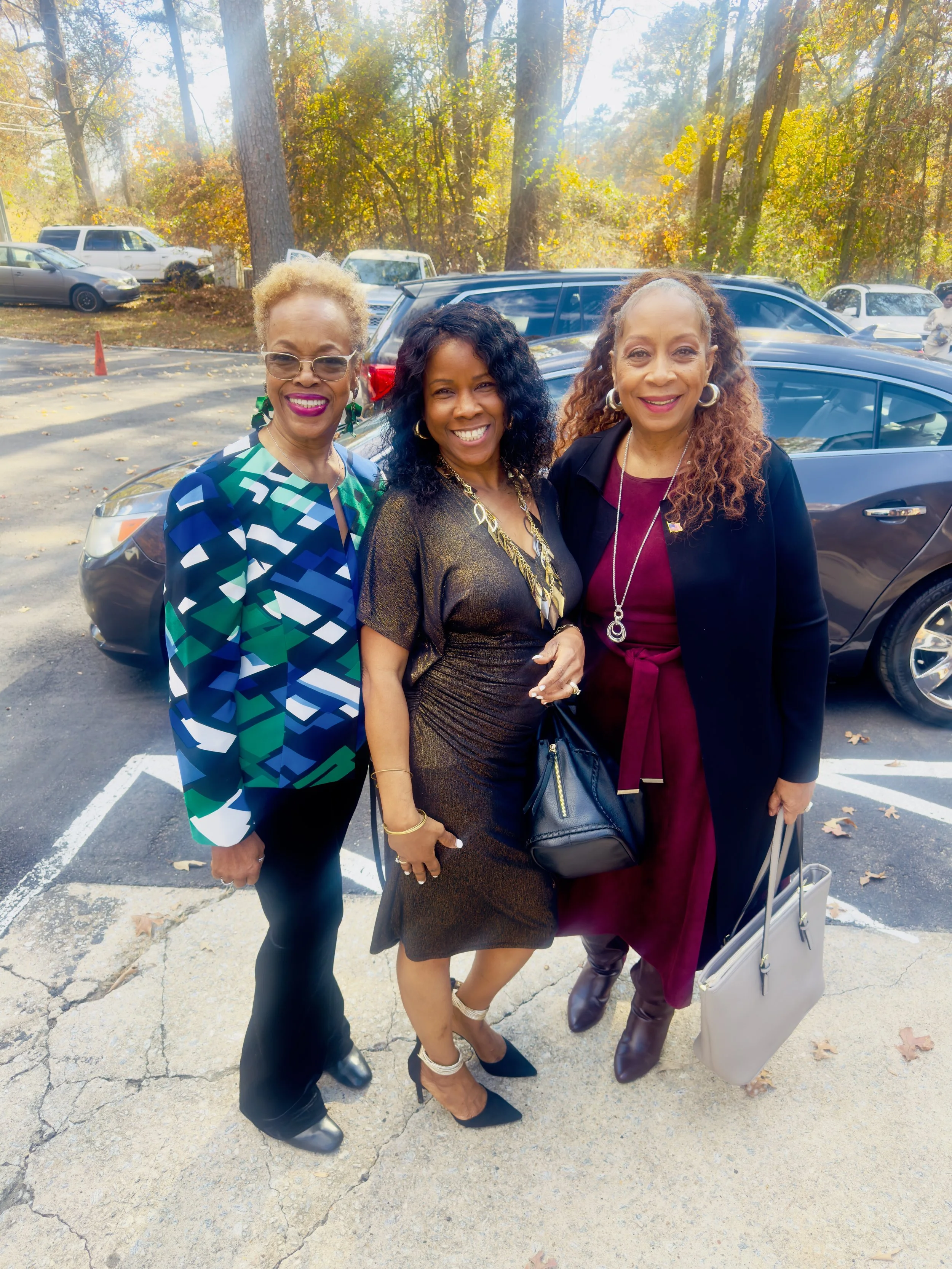 Three women standing together outdoors in a parking lot, smiling with trees and parked cars in the background, dressed in colorful and elegant clothing.