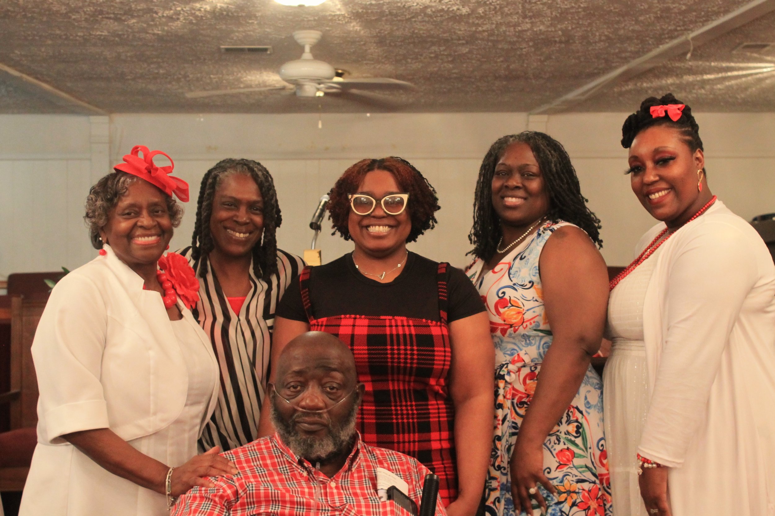 Six African American people, five women and one man, gather together indoors for a group photo. The women are dressed in colorful, patterned, and white outfits, with two wearing large red bows in their hair. The man, seated in front, has a beard and 