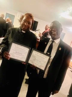 Two men in formal attire holding framed certificates at an indoor event, smiling for the photo.