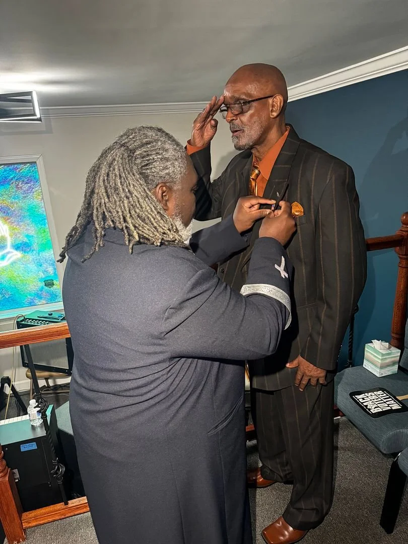 A woman pinning a medal on an older man in a striped suit during a ceremony in a room with a blue wall and a television screen.