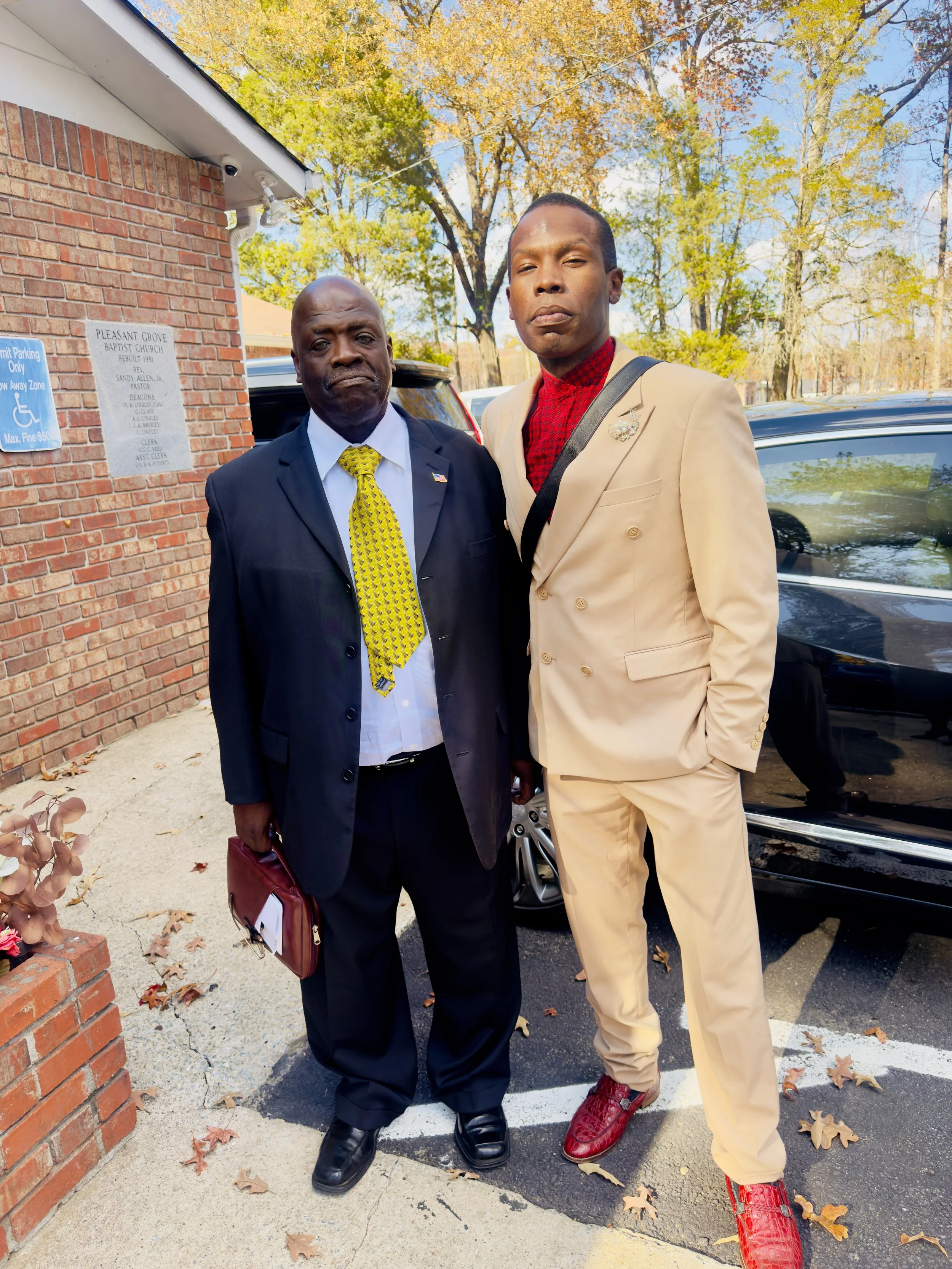 Two men standing outside near a brick building, one in a black suit with a yellow tie, and the other in a beige suit with red shoes, under trees with autumn foliage.