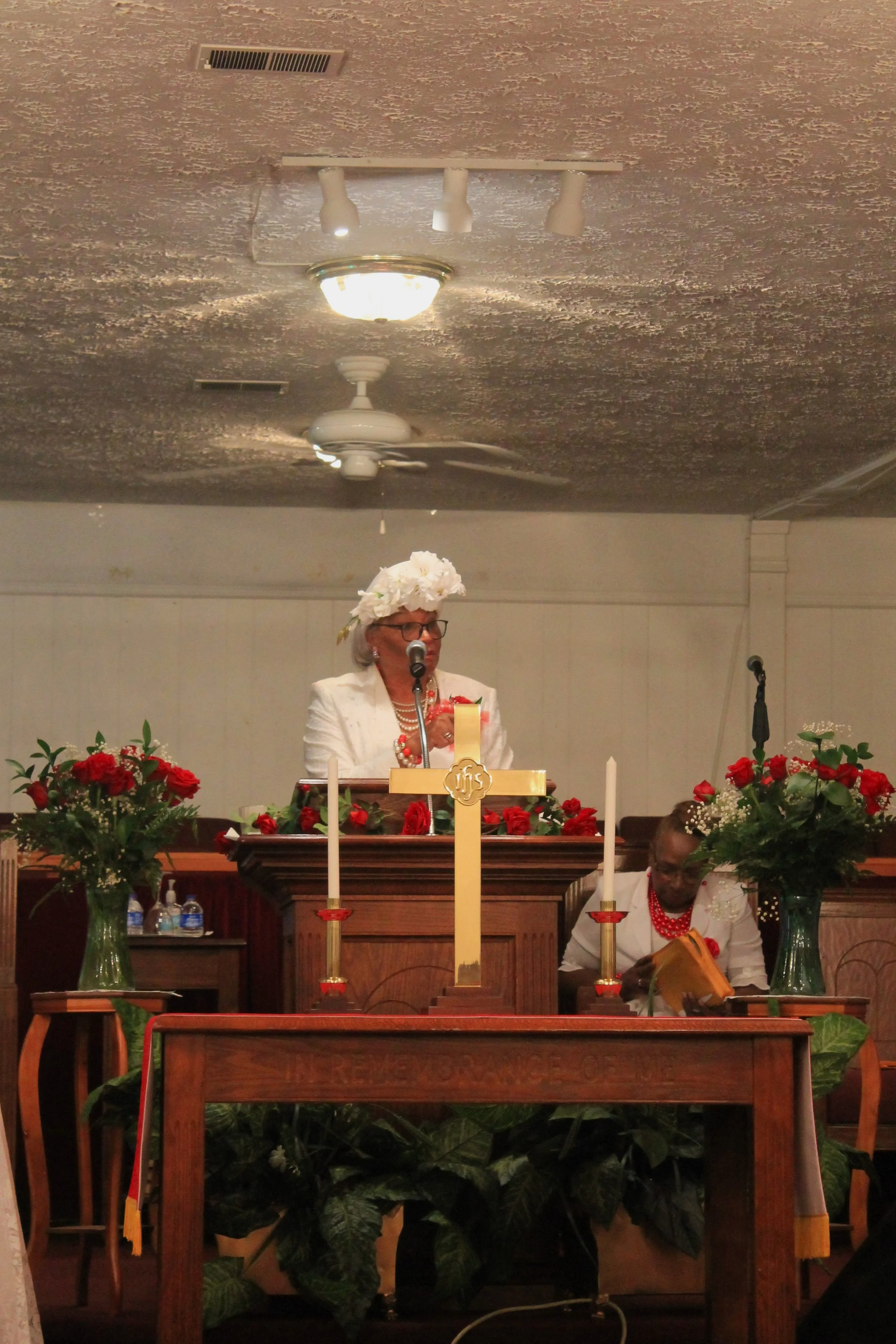 Woman standing at pulpit adorned with red roses, wearing white attire, a floral headpiece, and glasses, speaking into a microphone during a service in a church. Another woman seated to her right is reading a Bible.