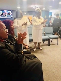 Group of women in a waiting area, some standing and others seated, with most wearing dark clothing and head coverings, engaged in conversation or using their phones.