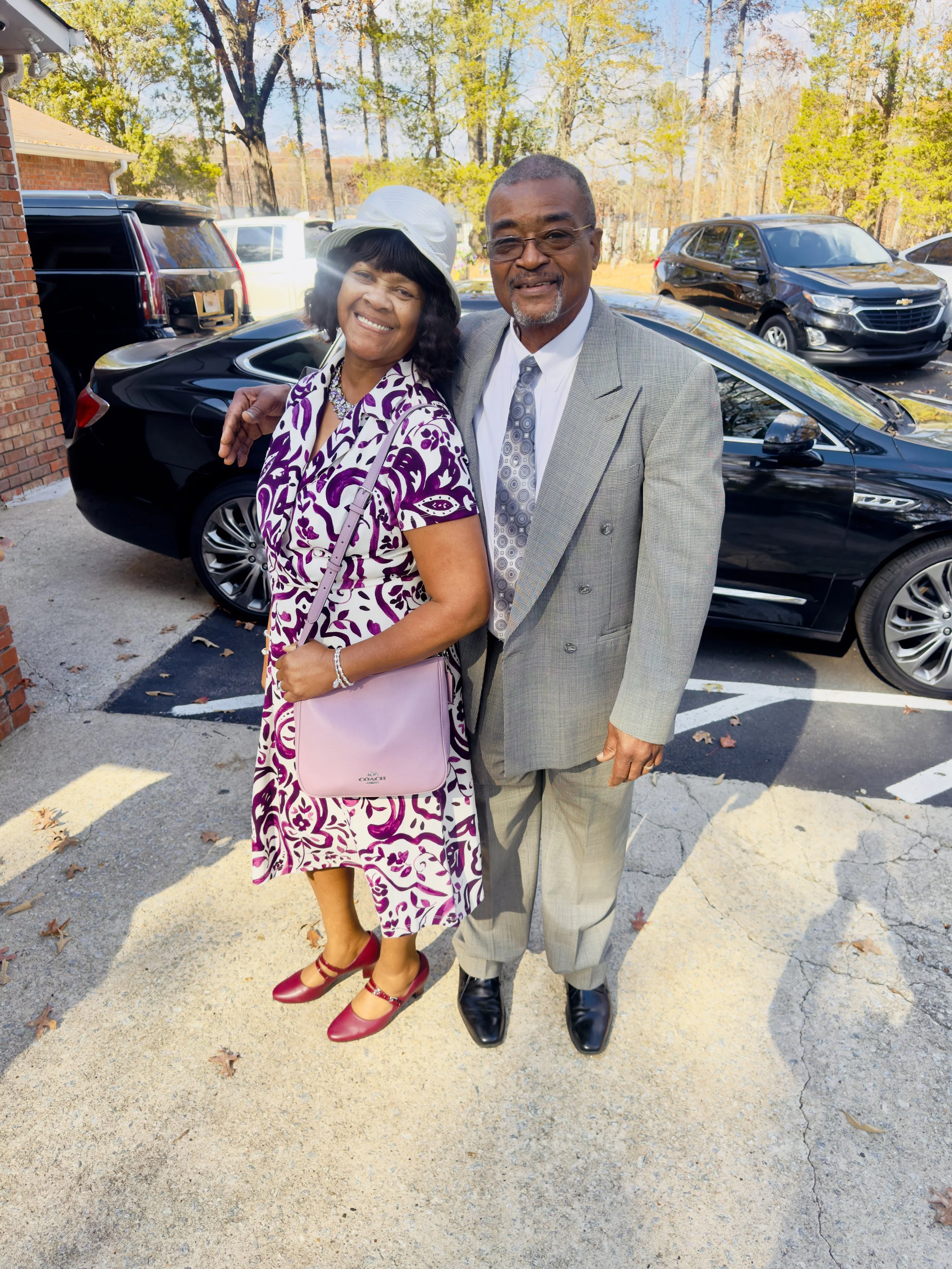 A smiling couple standing close together outdoors, dressed in elegant attire, with a row of parked cars and trees in the background.