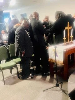 A group of people gathered around a table in a church, participating in a prayer or blessing ceremony, with an altar, cross, candles, and religious items present.