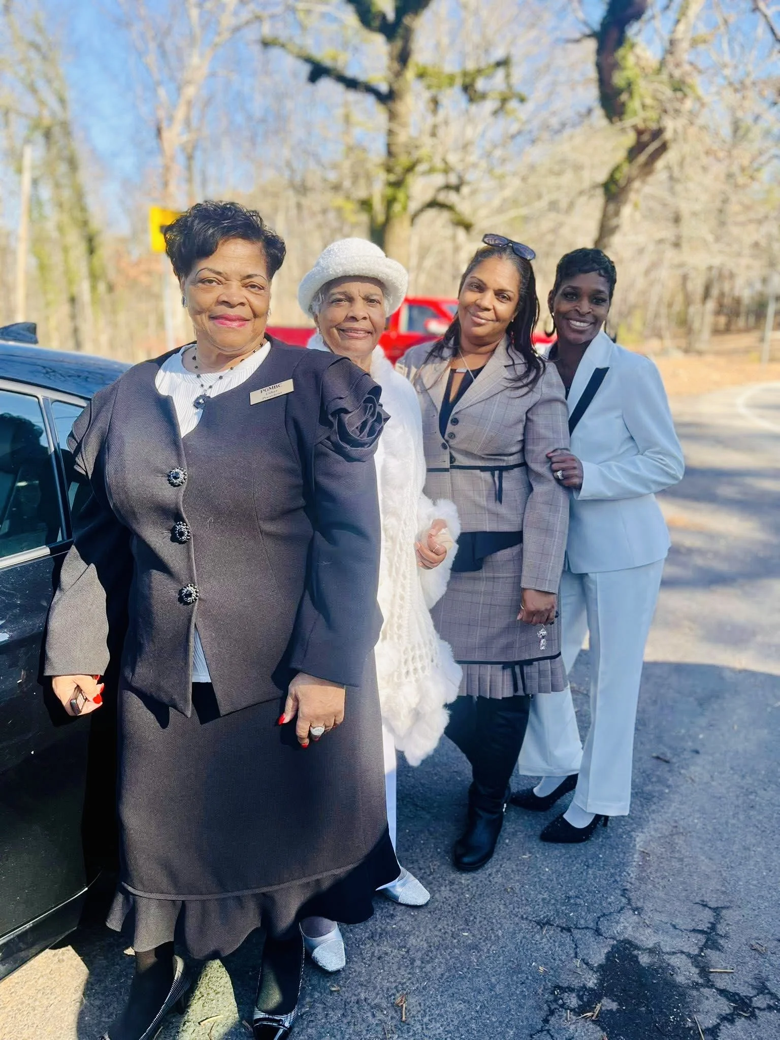Four women dressed elegantly, standing outdoors on a sunny day, with trees and a road in the background.