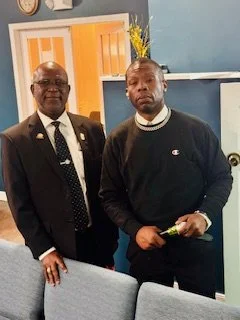 Two men standing indoors, one in a formal suit and the other in a black sweatshirt holding a small object, with a blue wall and a yellow floral decoration in the background.