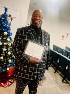 A man in a checkered suit holding a certificate, standing next to a decorated Christmas tree in a room with blue chairs and holiday decorations.