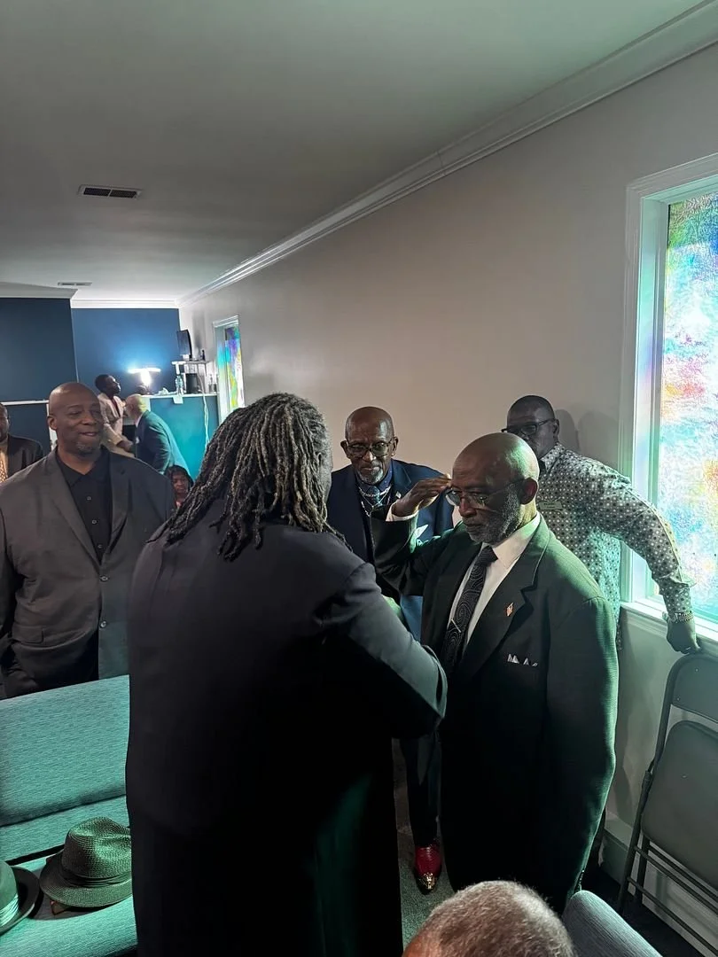 A group of men in formal attire gathered in a room near a window, engaged in conversation, with colorful stained glass and a green couch in the background.
