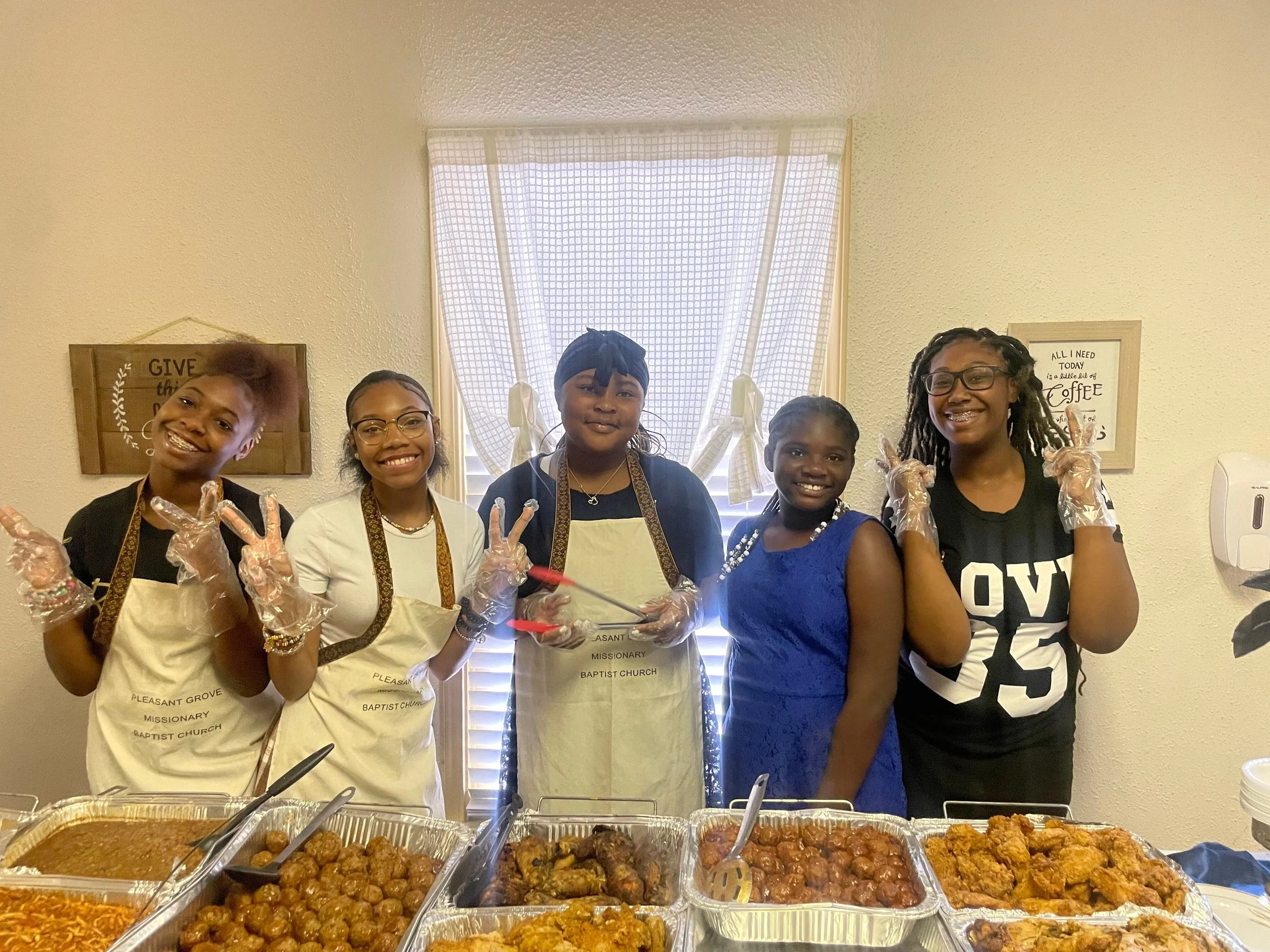 Group of five women standing behind a table of food, smiling and making peace signs. They are wearing gloves and aprons, with a window and decorative signs on the wall in the background.