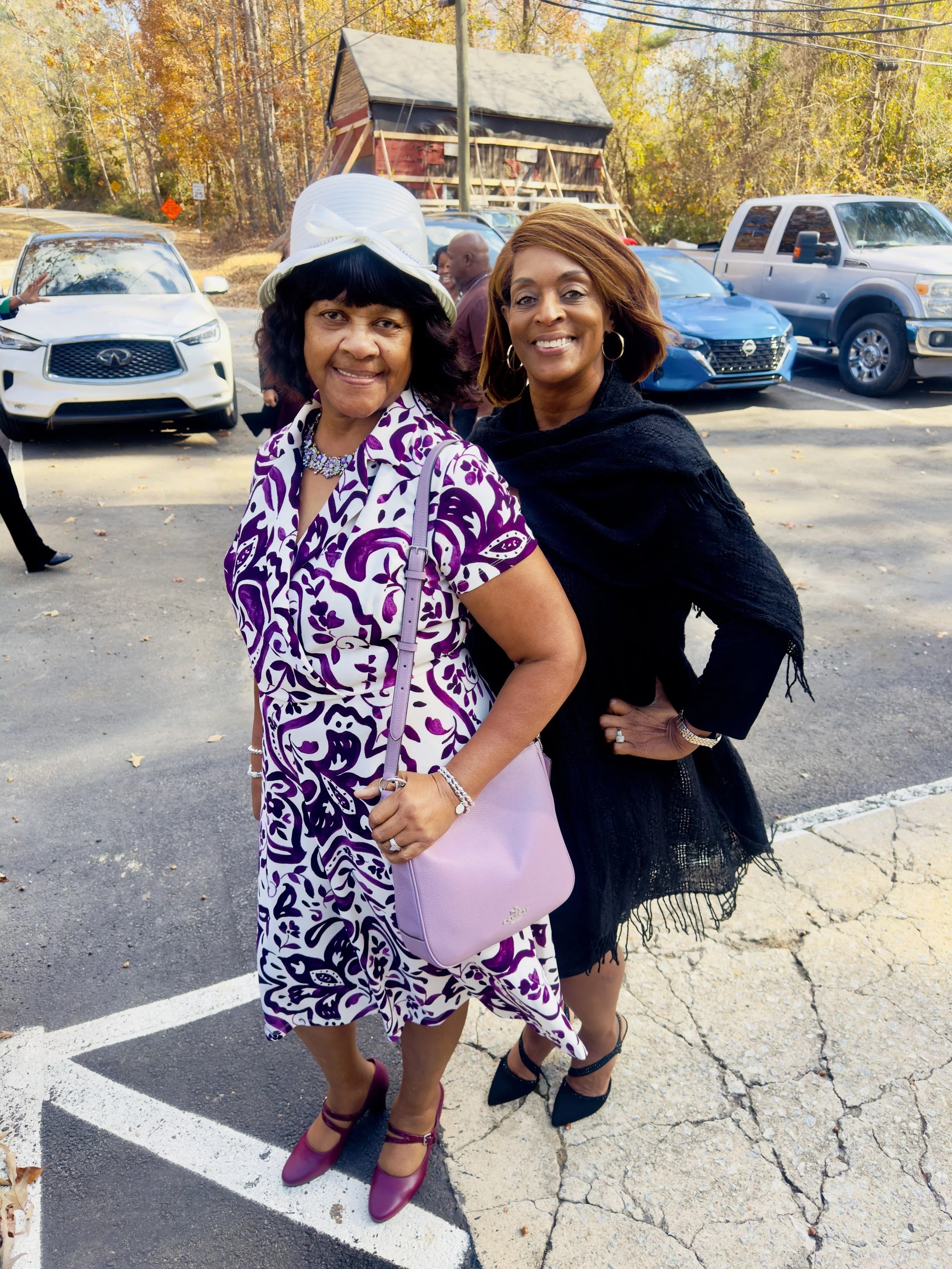 Two women standing outdoors in a parking lot, smiling at the camera. One woman is wearing a purple and white patterned dress with a white hat, and the other is dressed in a black outfit with a shawl. There are cars and trees with fall foliage in the 