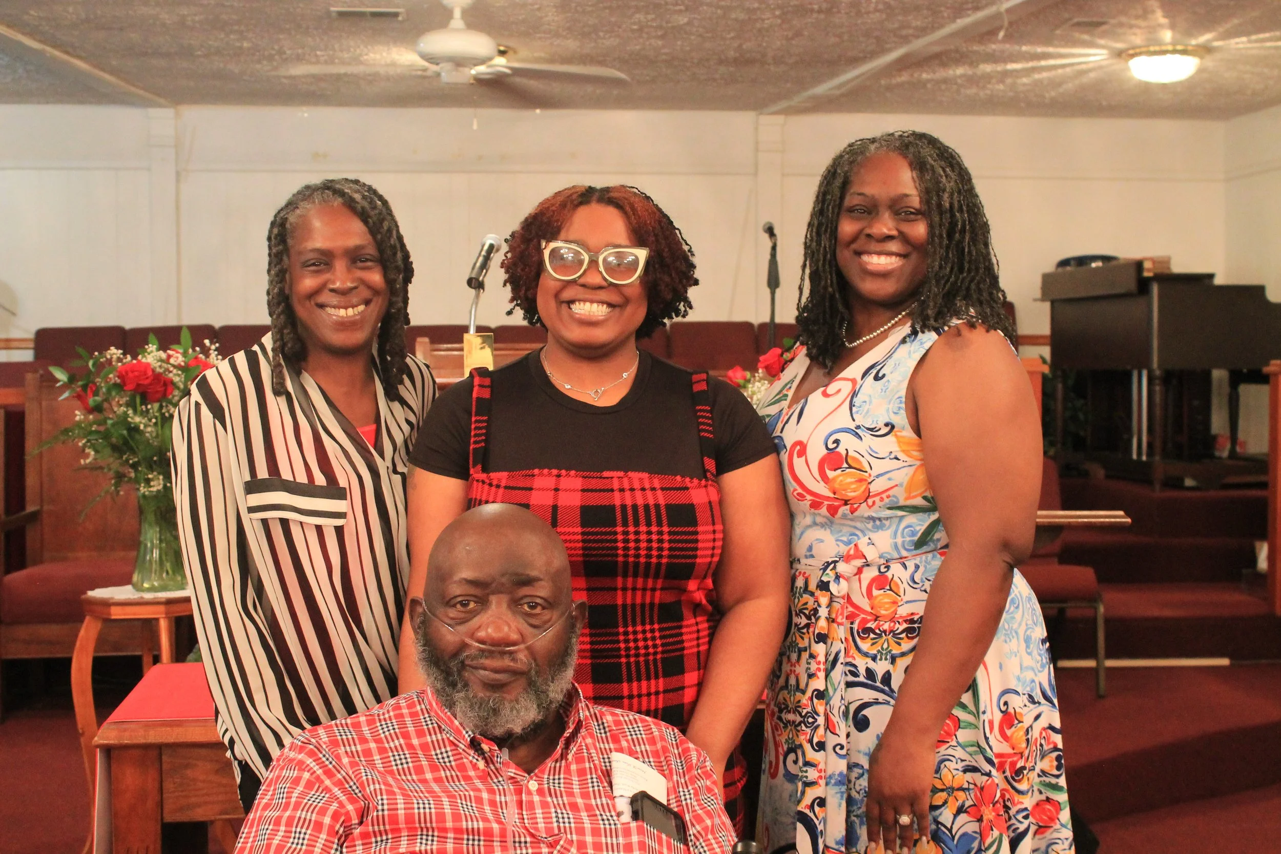 Four people, three women and one man, smiling in a church or community hall setting. The man is seated in the front wearing a red plaid shirt, and the women are standing behind him. Two women are wearing patterned dresses, and one has glasses. Flower