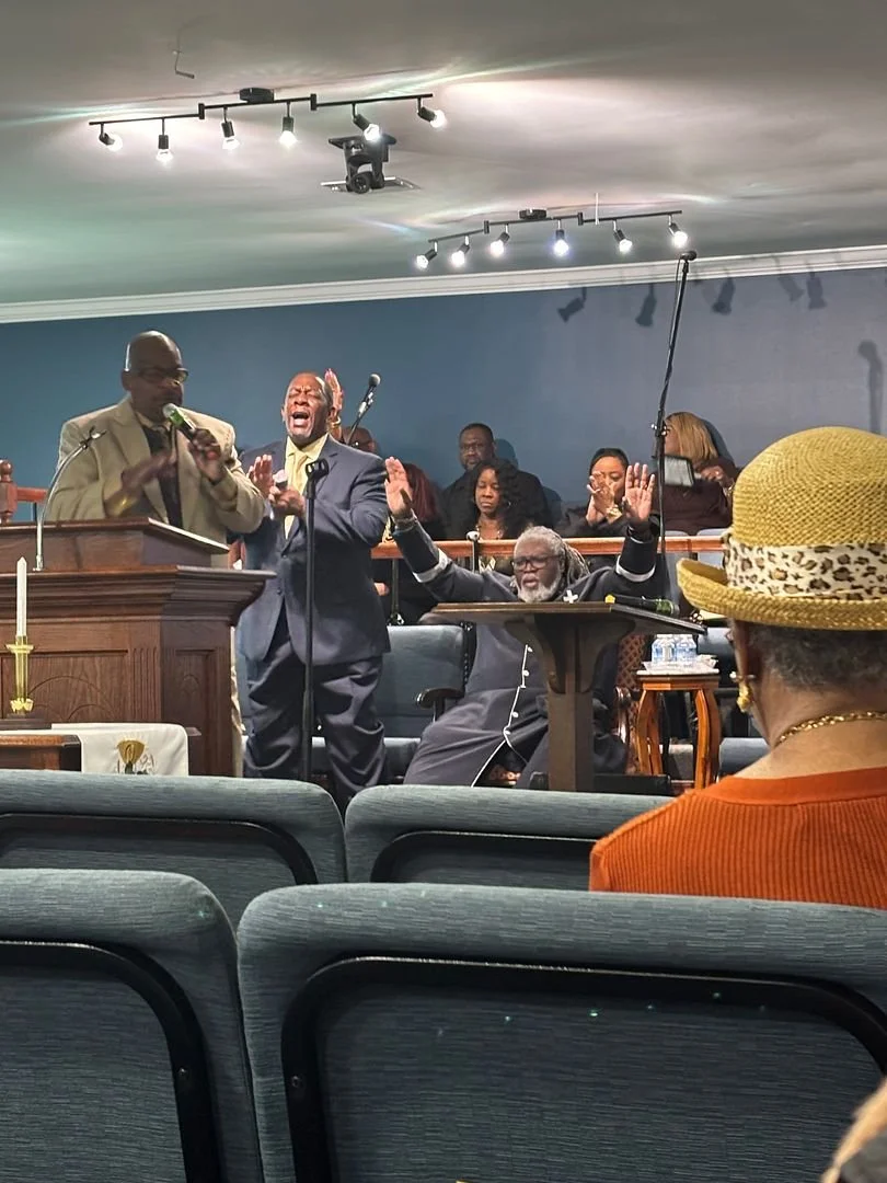 A church service with people singing and clapping, including a man at the pulpit, a seated man with raised hands, and others singing in the background. A woman wearing a straw hat is visible in the foreground.