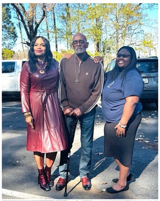 Three people standing outdoors in a parking lot with trees and cars in the background. An elderly man in the center with a cane, flanked by women on each side, all smiling.