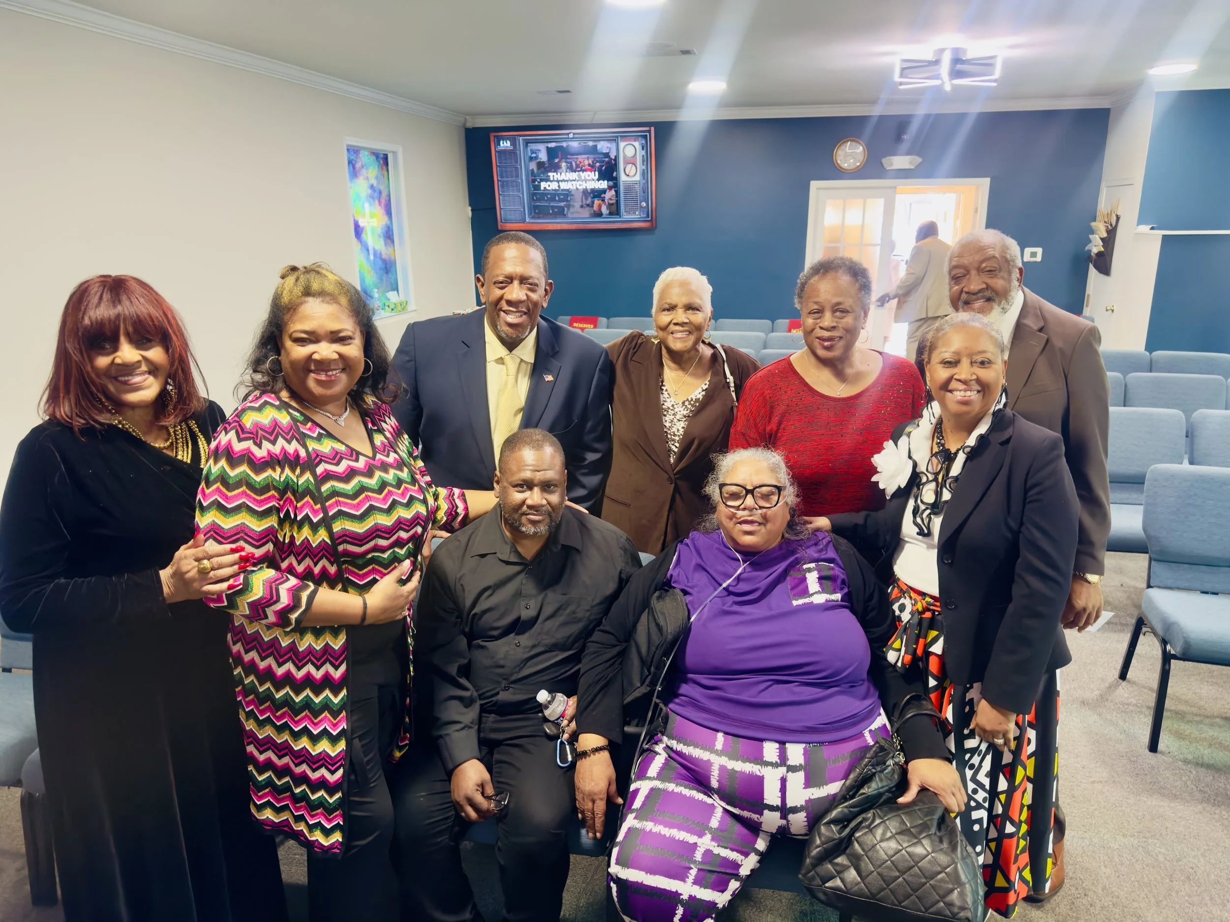 A group of eleven people, including men and women of diverse ages, posing together indoors with a church or community center setting in the background.
