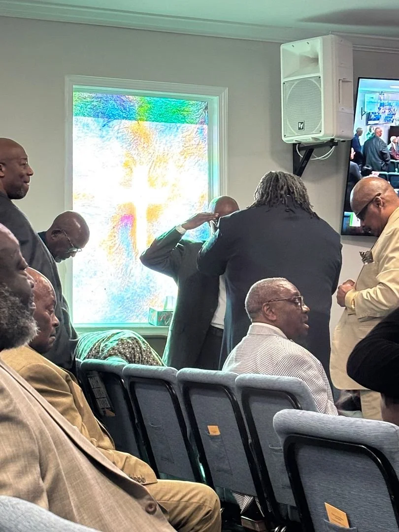 Group of men in a meeting, some seated and some standing, in a conference room with a bright window and a wall-mounted TV screen showing a video conference.