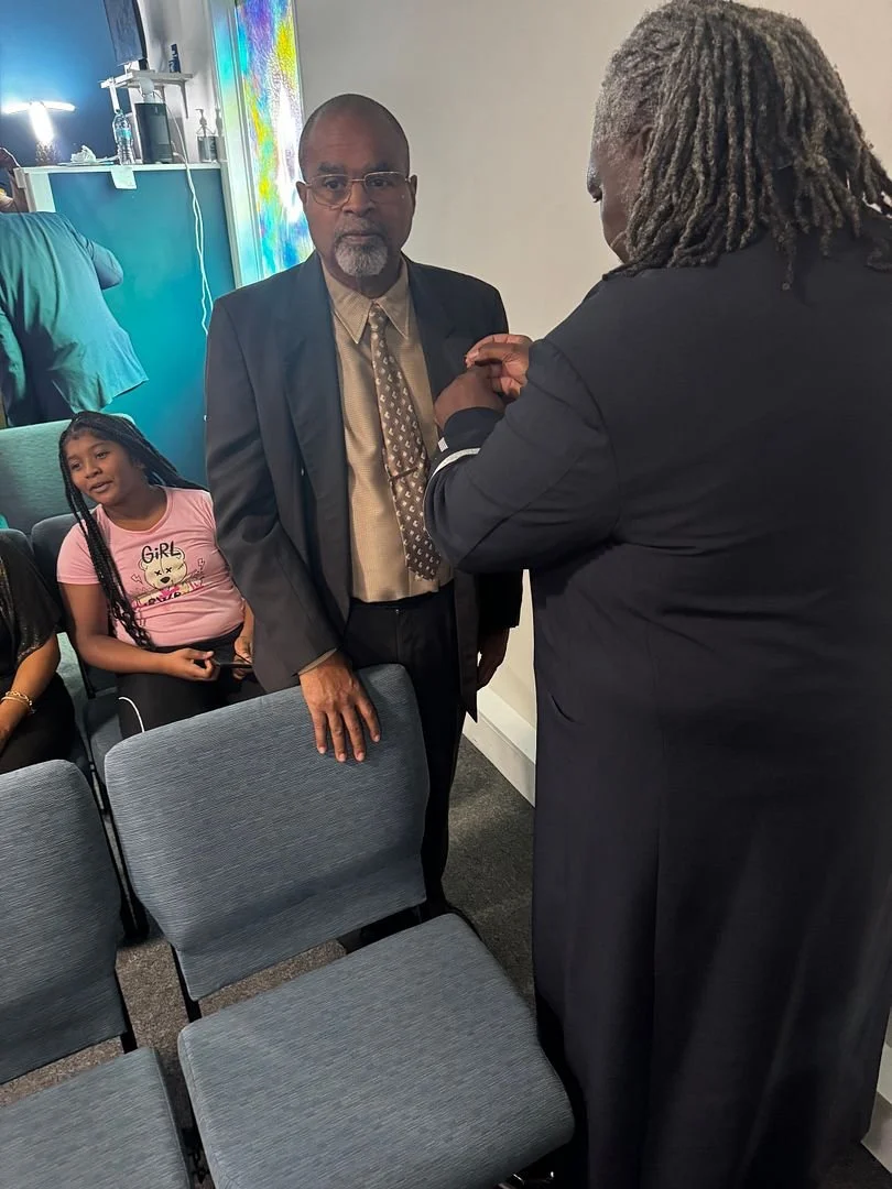 A man in a suit with glasses and a beard stands in a room, being officiated at by another man with long gray dreadlocks who is pinning or adjusting a small item on his suit. A young girl with braided hair, wearing a pink T-shirt with a cartoon cat an