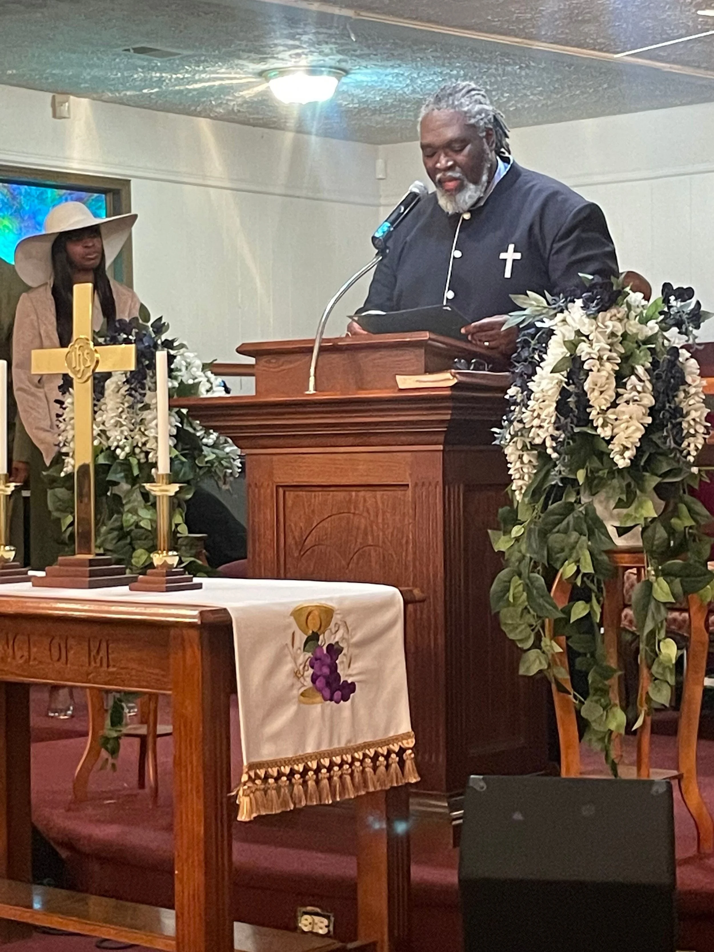 A man in a black clergy shirt with a white cross on the chest is standing at a wooden pulpit, reading from a black book during a church service. Behind him, a woman in a wide-brimmed hat stands near a stained glass window. The altar is decorated with