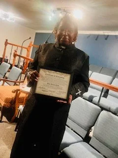 Woman in black holding a framed certificate, standing in a room with blue walls and gray chairs.