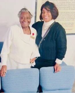 Two women standing indoors, smiling, with chairs and a framed document or sign behind them.