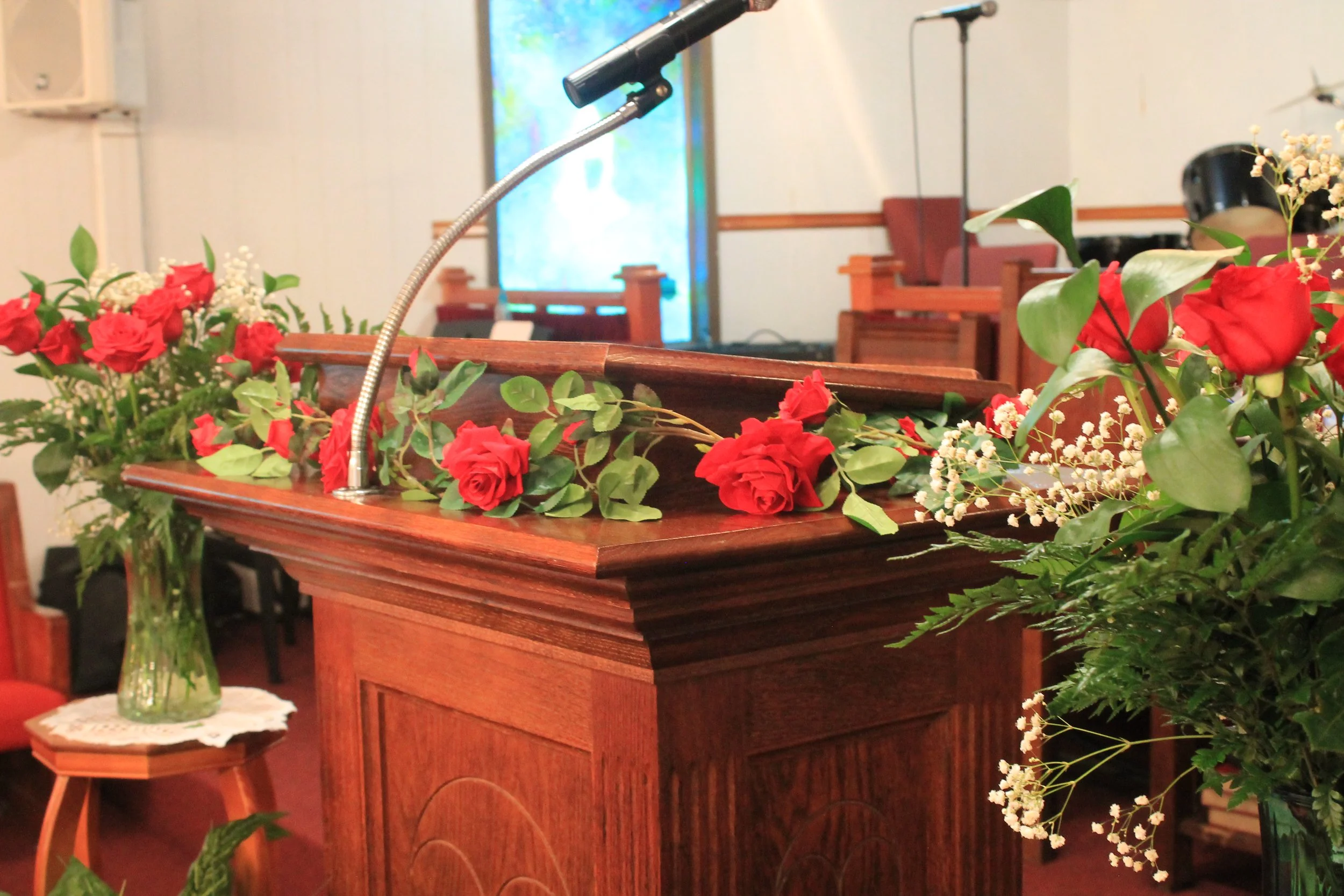 A wooden podium decorated with red roses and green leaves in a church interior, with more floral arrangements and a microphone.