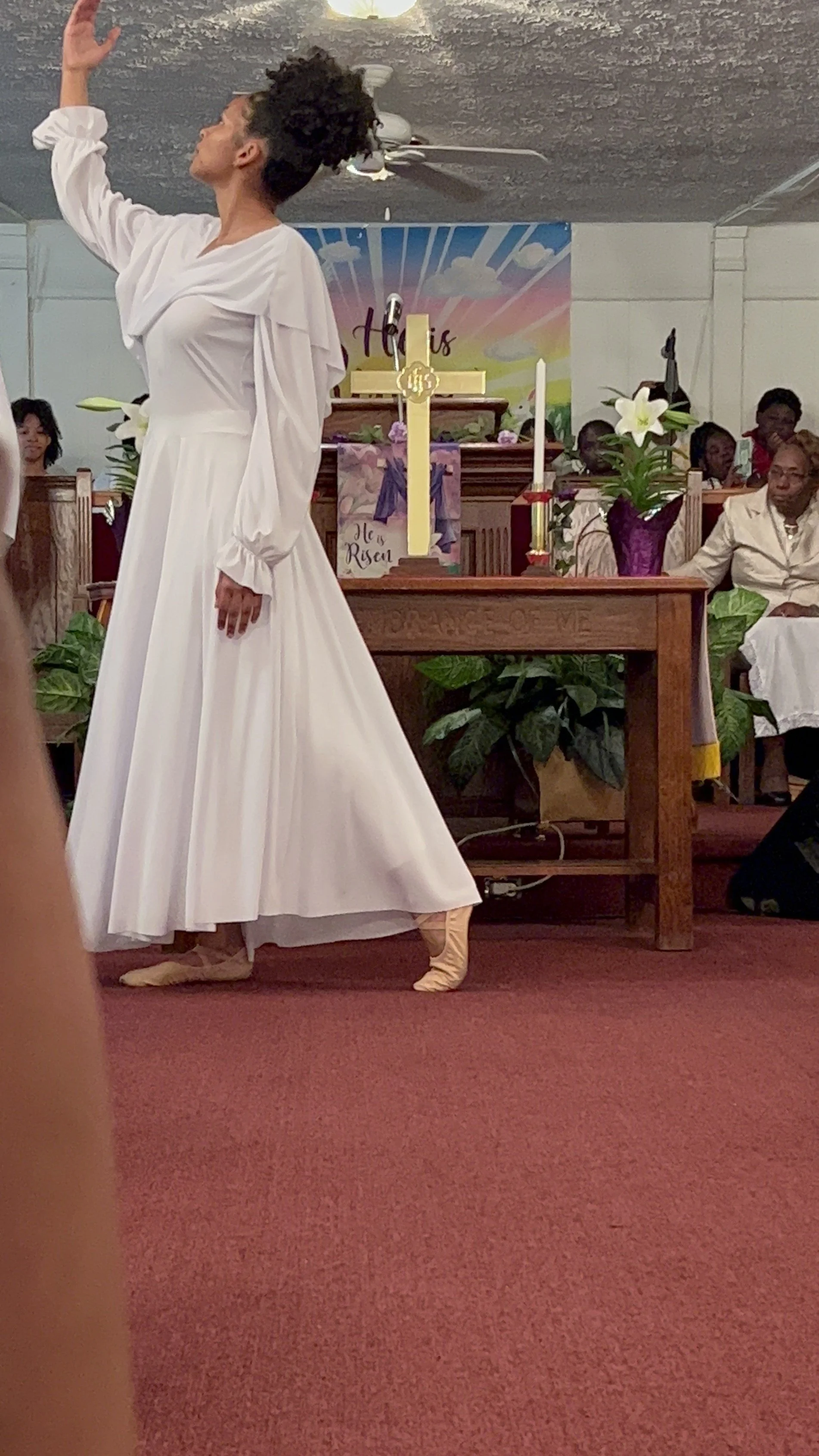 A woman in a white dress and ballet slippers dancing at the front of a church, with people seated in pews behind her, and religious decorations including a cross, candle, and flowers on the altar.